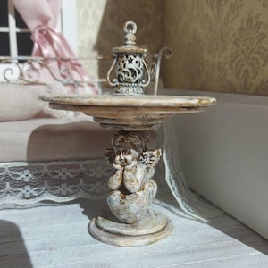 Könnte beinhalten: Ein Miniaturtisch im antiken Stil mit einem verwitterten, cremefarbenen Finish. Der Tisch hat eine runde Platte und wird von einer Cherubim-Figur getragen. Auf dem Tisch steht eine kleine, verzierte Laterne, die zur Vintage-Ästhetik beiträgt.