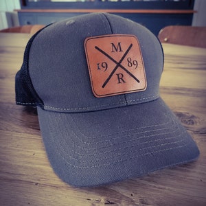 Puede incluir: Una gorra de camionero gris y negra con un parche de cuero marrón que dice "M 19 89 R" en un diseño de cruz.