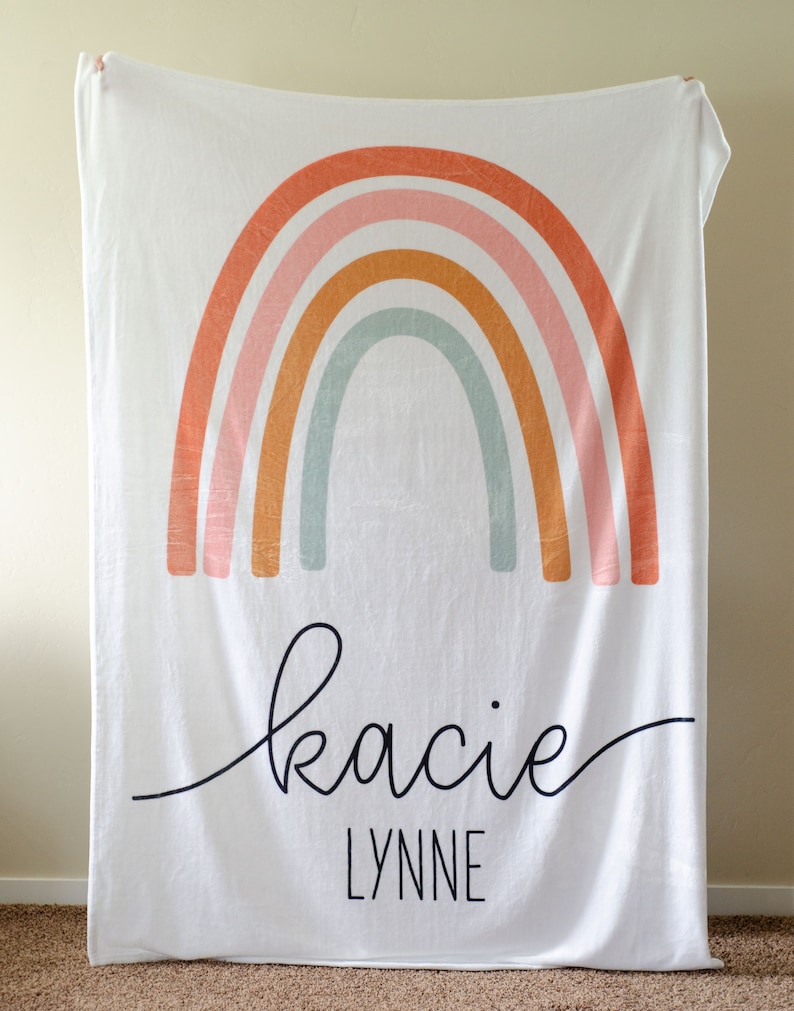 May include: White fleece blanket with a rainbow design in shades of orange, pink, and green. The blanket has the name "Kacie Lynne" written in black cursive script.