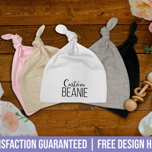 May include: A collection of baby beanies in various colors: pink, beige, white, gray, and black. The white beanie has the text "Custom BEANIE" printed on it. The beanies are displayed on a wooden surface, with a floral blanket and baby toys in the background.