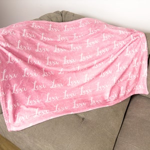 May include: A pink fleece blanket with the name "Lexi" printed in white cursive script all over the blanket.