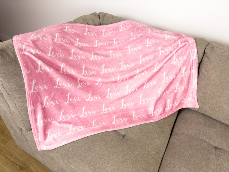May include: A pink fleece blanket with the name "Lexi" printed in white cursive script all over the blanket.