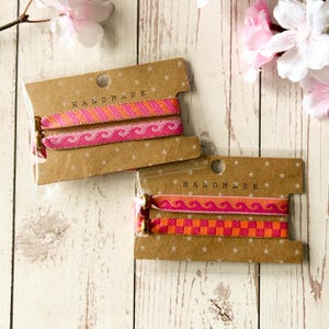 May include: Two sets of two elastic hair ties wrapped around brown cardboard cards labelled HANDMADE. The hair ties feature pink and orange woven patterns, including waves and checkered designs, secured with small metallic gold-toned beads.