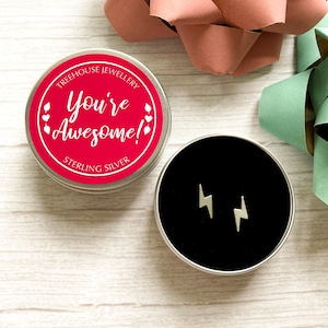 May include: Sterling silver lightning bolt stud earrings presented in an open tin with a black interior. A red tin with the text "You're Awesome!" and "Sterling Silver" is also visible. The background features gift bows.