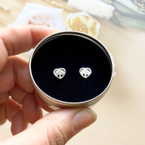 May include: A pair of silver heart-shaped earrings with a tree design inside the heart. The earrings are in a small round tin box with a black interior.
