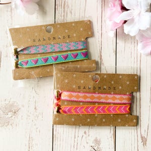 May include: Two sets of woven fabric bracelets on brown cardboard display cards labelled HANDMADE. The top set features a pink and teal chevron pattern and a teal band with pink hearts. The bottom set has a pink and orange diamond pattern and a pink and orange chevron pattern, all secured with gold-toned metal beads.