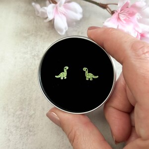 May include: A pair of small stud earrings shaped like long-necked dinosaurs. Each earring is bright lime green with a textured surface and a single black dot for the eye. The earrings are displayed inside a small, round, silver-coloured metal tin.