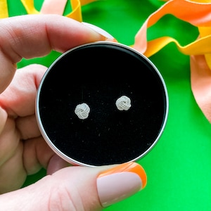 May include: A small round black box with a lid open, revealing two silver earrings. The earrings are in the shape of a knot. The box is sitting on a hand with a light pink nail polish.