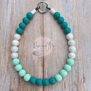 May include: A teal, white, and mint green beaded necklace with a silver clasp. The necklace is made of round beads and is displayed on a wooden surface. The brand name "Rose and Ale" is visible.