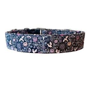 May include: A dog collar with a black plastic buckle and silver-colored metal hardware. The collar features a dark blue fabric with a floral pattern in shades of pink, purple, and white. The design includes stylized birds and flowers.