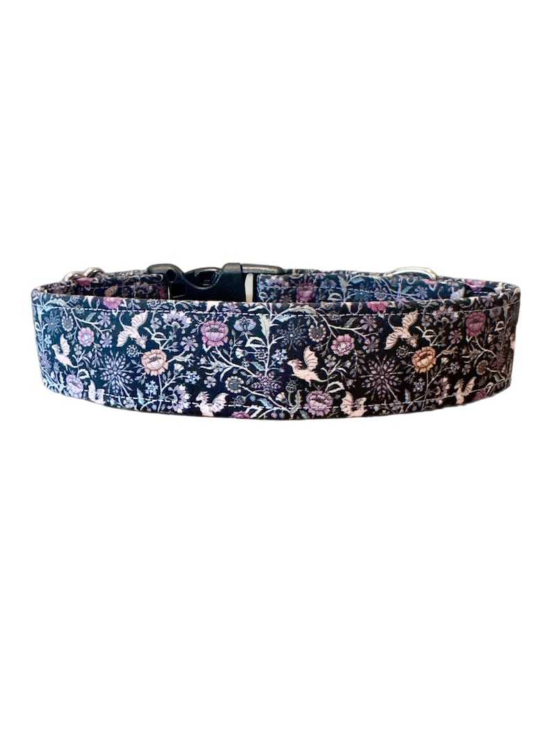 May include: A dog collar with a black plastic buckle and silver-colored metal hardware. The collar features a dark blue fabric with a floral pattern in shades of pink, purple, and white. The design includes stylized birds and flowers.