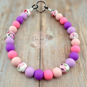May include: A pink and purple silicone beaded dog collar with a floral print on some of the beads. The collar has a silver clasp and is attached to a silver ring.