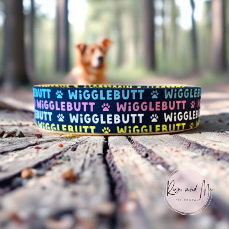 Op de afbeelding: Een marineblauwe hondenriem met de tekst "WIGGLEBUTT" in regenboogkleuren en pootafdrukken. De halsband heeft een gele rand en staat tegen een wazige achtergrond van een hond en een bosrijk gebied.