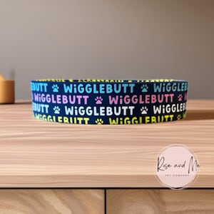 Op de afbeelding: Een marineblauwe voerbak voor huisdieren met de woorden "Wigglebutt" in een herhalend patroon van kleurrijke tekst en pootafdrukken. De kom staat op een houten oppervlak.