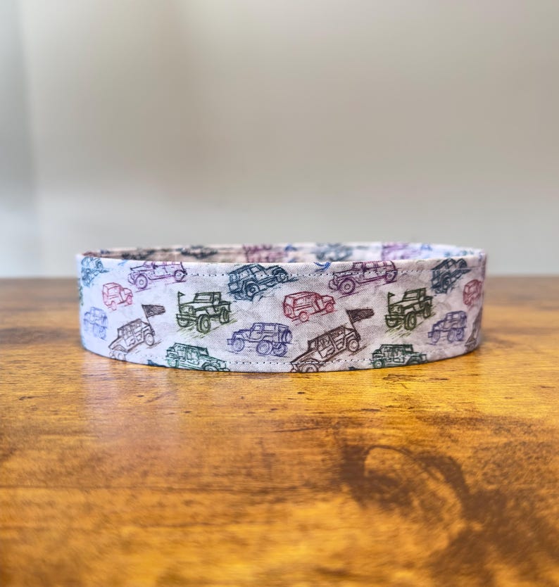 May include: A white fabric collar with a repeating pattern of colorful cartoon off-road vehicles. The vehicles are drawn in a simple style and are outlined in black.