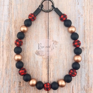 May include: A beaded pet collar featuring a mix of black, gold, and red buffalo plaid beads. The collar has a black clasp and connectors, and the text "Rose and Me PET COMPANY" is visible.