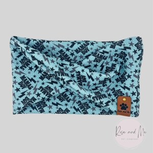 May include: A light blue pet bandana with a repeating pattern of black stars, lightning bolts, and the text "Mamas Boy." A small brown leather tag with a paw print logo is attached. The bandana is designed for pets.