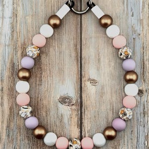 K&ouml;nnte beinhalten: Eine Perlenkette mit einem runden silberfarbenen Metallverschluss am oberen Ende. Die Perlen wechseln sich ab zwischen metallischem Bronze, mattem Wei&szlig;, zartem Rosa, hellem Violett und gemusterten Perlen mit kleinen Geistern, K&uuml;rbissen und Spinnennetzen.