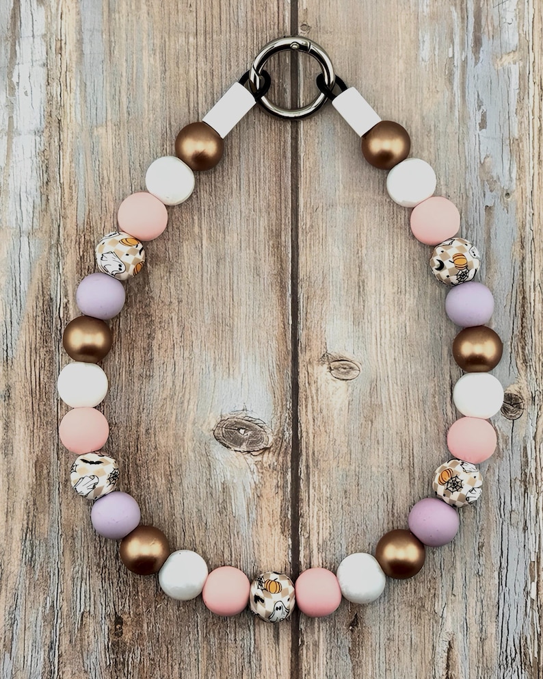 K&ouml;nnte beinhalten: Eine Perlenkette mit einem runden silberfarbenen Metallverschluss am oberen Ende. Die Perlen wechseln sich ab zwischen metallischem Bronze, mattem Wei&szlig;, zartem Rosa, hellem Violett und gemusterten Perlen mit kleinen Geistern, K&uuml;rbissen und Spinnennetzen.