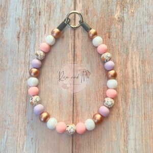 K&ouml;nnte beinhalten: Ein kreisf&ouml;rmiges Haustierhalsband mit einer Abfolge von kugelf&ouml;rmigen Perlen in Ros&eacute;gold, mattem Wei&szlig;, zartem Rosa und Lavendel, erg&auml;nzt durch gemusterte Perlen mit Blumenmotiven und einem silbernen Metallring als Verschluss.