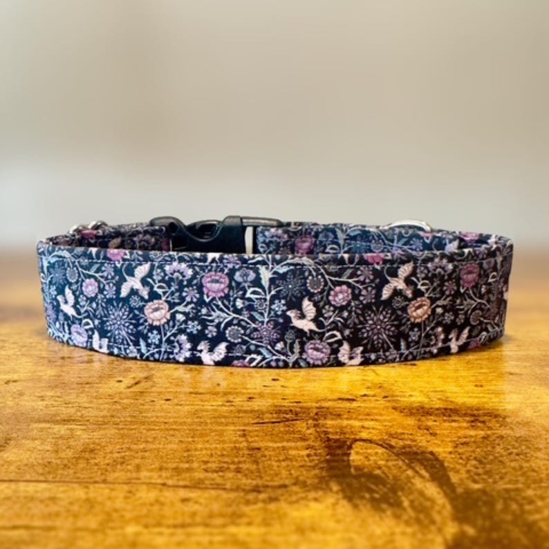 May include: A dog collar with a floral pattern in navy blue, pink, and white. The collar has a black plastic buckle and metal hardware. The design features flowers and birds.