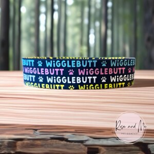 Op de afbeelding: Een marineblauwe hondenvoerbak met het woord "Wigglebutt" in een herhalend patroon van kleurrijke tekst en pootafdrukken. De bak staat op een houten oppervlak met een vage bosachtergrond.