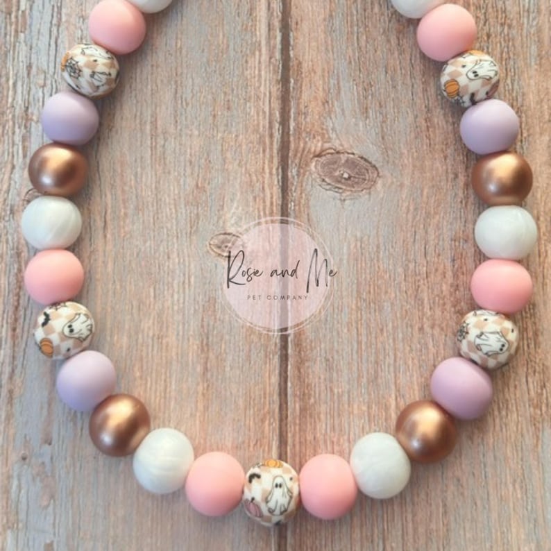 K&ouml;nnte beinhalten: Eine Perlenkette mit einem sich wiederholenden Muster aus runden Perlen in mattem Rosa, Lavendel, Wei&szlig; und metallischem Kupfer, durchsetzt mit gemusterten Perlen, die kleine wei&szlig;e Geister und orangefarbene K&uuml;rbisse auf kariertem Grund zeigen.