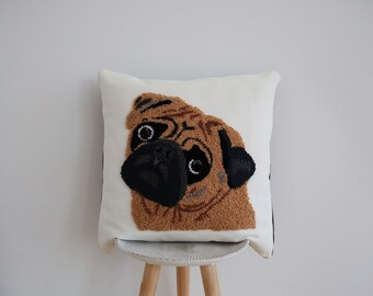 May include: White pillow with a brown and black pug face embroidered on it. The pillow is sitting on a wooden stool.