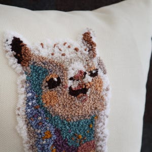 May include: A cream-colored throw pillow featuring a textured punch needle embroidery of a llama. The llama is depicted with a multi-colored coat in shades of brown, white, teal, and purple, with dark eyes and a smiling expression on its face.