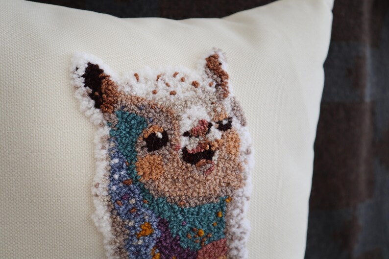 May include: A cream-colored throw pillow featuring a textured punch needle embroidery of a llama. The llama is depicted with a multi-colored coat in shades of brown, white, teal, and purple, with dark eyes and a smiling expression on its face.