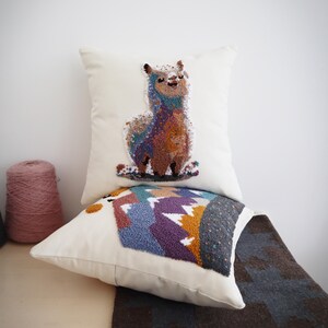 May include: Two square off-white throw pillows featuring textured punch needle embroidery. The top pillow displays a colorful llama, while the bottom pillow depicts a mountain landscape with purple, blue, and gold peaks against a dark gray sky.