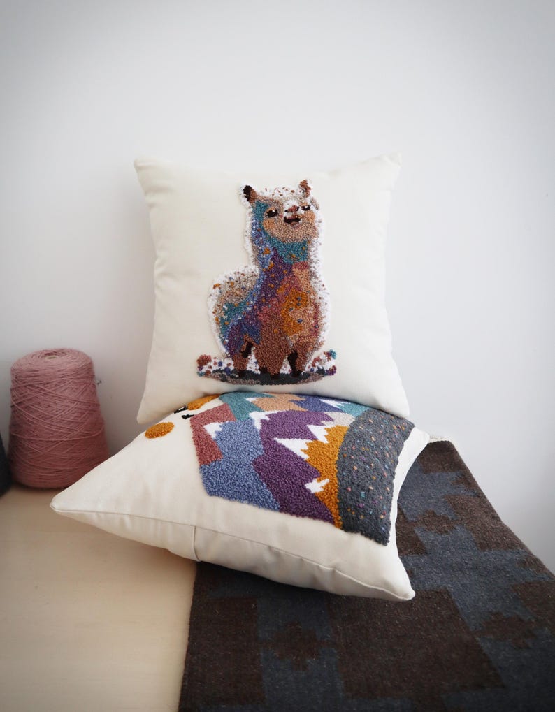 May include: Two square off-white throw pillows featuring textured punch needle embroidery. The top pillow displays a colorful llama, while the bottom pillow depicts a mountain landscape with purple, blue, and gold peaks against a dark gray sky.