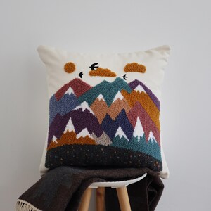 May include: A square throw pillow featuring a punch needle mountain landscape design. The scene includes layered peaks in shades of purple, teal, brown, and white, with a mustard yellow sun, two textured clouds, and three black birds against a cream background.
