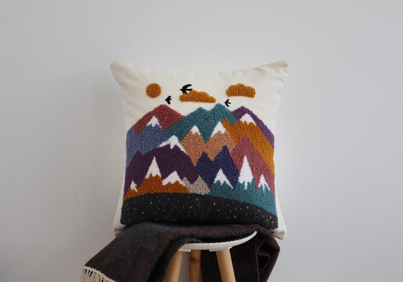 May include: A square throw pillow featuring a punch needle mountain landscape design. The scene includes layered peaks in shades of purple, teal, brown, and white, with a mustard yellow sun, two textured clouds, and three black birds against a cream background.