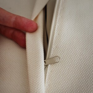 May include: An off-white canvas fabric cushion cover featuring a concealed zipper closure. The textured material has a visible weave pattern, and the metal zipper pull is positioned along the seam, allowing for easy access to the interior.