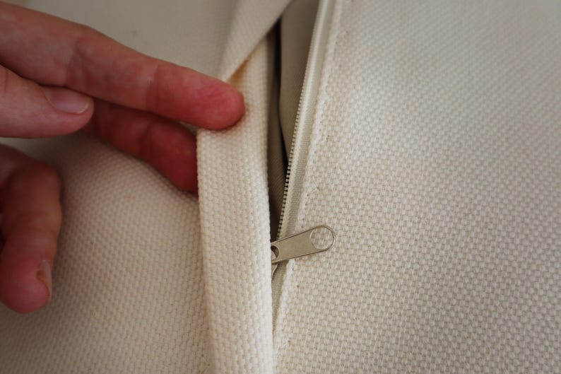 May include: An off-white canvas fabric cushion cover featuring a concealed zipper closure. The textured material has a visible weave pattern, and the metal zipper pull is positioned along the seam, allowing for easy access to the interior.
