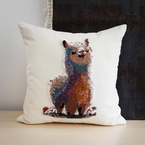 May include: A square off-white throw pillow featuring a central punch needle embroidery design of a standing llama. The llama is rendered in a multi-colored gradient of blue, purple, orange, and brown yarn, creating a textured, speckled appearance.