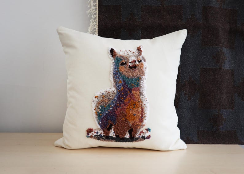 May include: A square off-white throw pillow featuring a central punch needle embroidery design of a standing llama. The llama is rendered in a multi-colored gradient of blue, purple, orange, and brown yarn, creating a textured, speckled appearance.