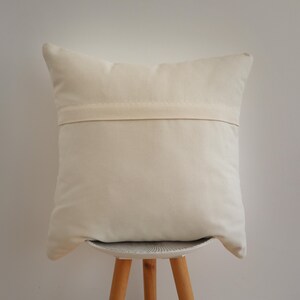 May include: A square off-white throw pillow cover featuring a horizontal fabric flap across the center, which conceals a zipper closure. The fabric has a plain, solid-colored texture and a neutral, light cream tone throughout the entire surface.