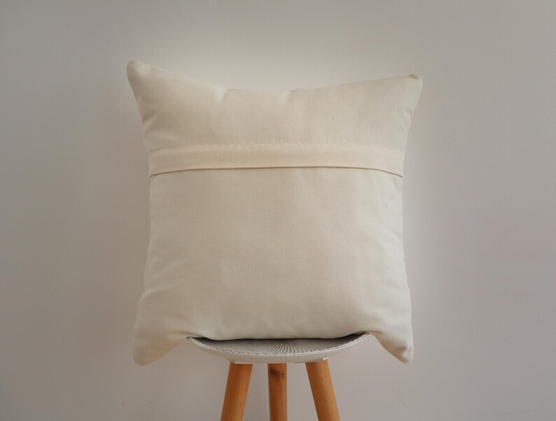 May include: A square off-white throw pillow cover featuring a horizontal fabric flap across the center, which conceals a zipper closure. The fabric has a plain, solid-colored texture and a neutral, light cream tone throughout the entire surface.