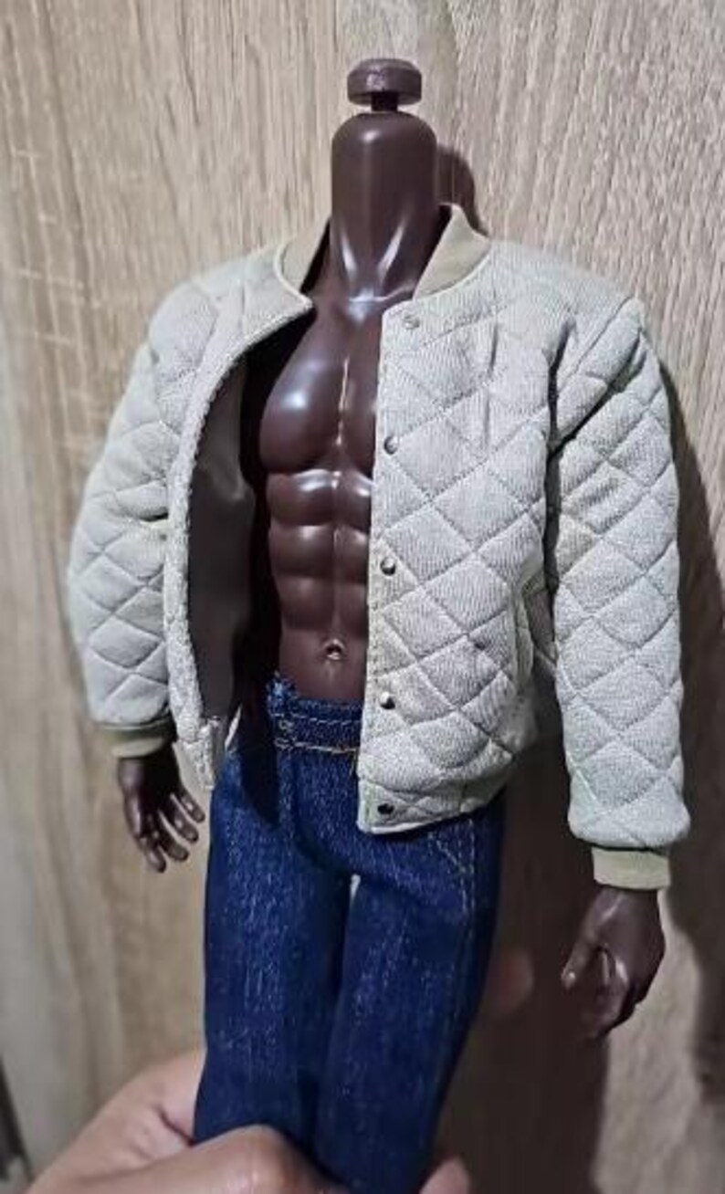 May include: A dark-skinned articulated doll figure wearing a beige quilted bomber jacket with a snap-button front closure and dark blue denim jeans. The jacket features a textured diamond-patterned fabric and ribbed cuffs, worn open over the torso.