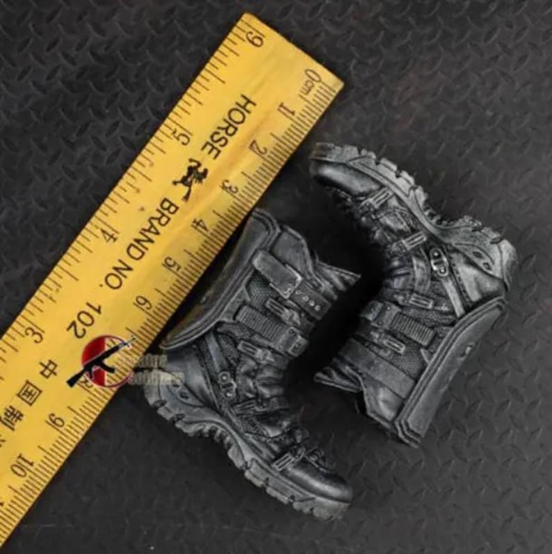 May include: A pair of miniature black combat boots with multiple straps and buckles. A yellow ruler with black markings and the text "HORSE BRAND NO. 102" is in the background. The boots are approximately 3 inches in length.