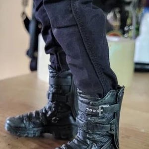 May include: Black combat boots with multiple buckles and straps. The boots have a rugged, textured appearance and are worn with black pants. The boots appear to be made of leather or a similar material, with visible stitching and detailing.