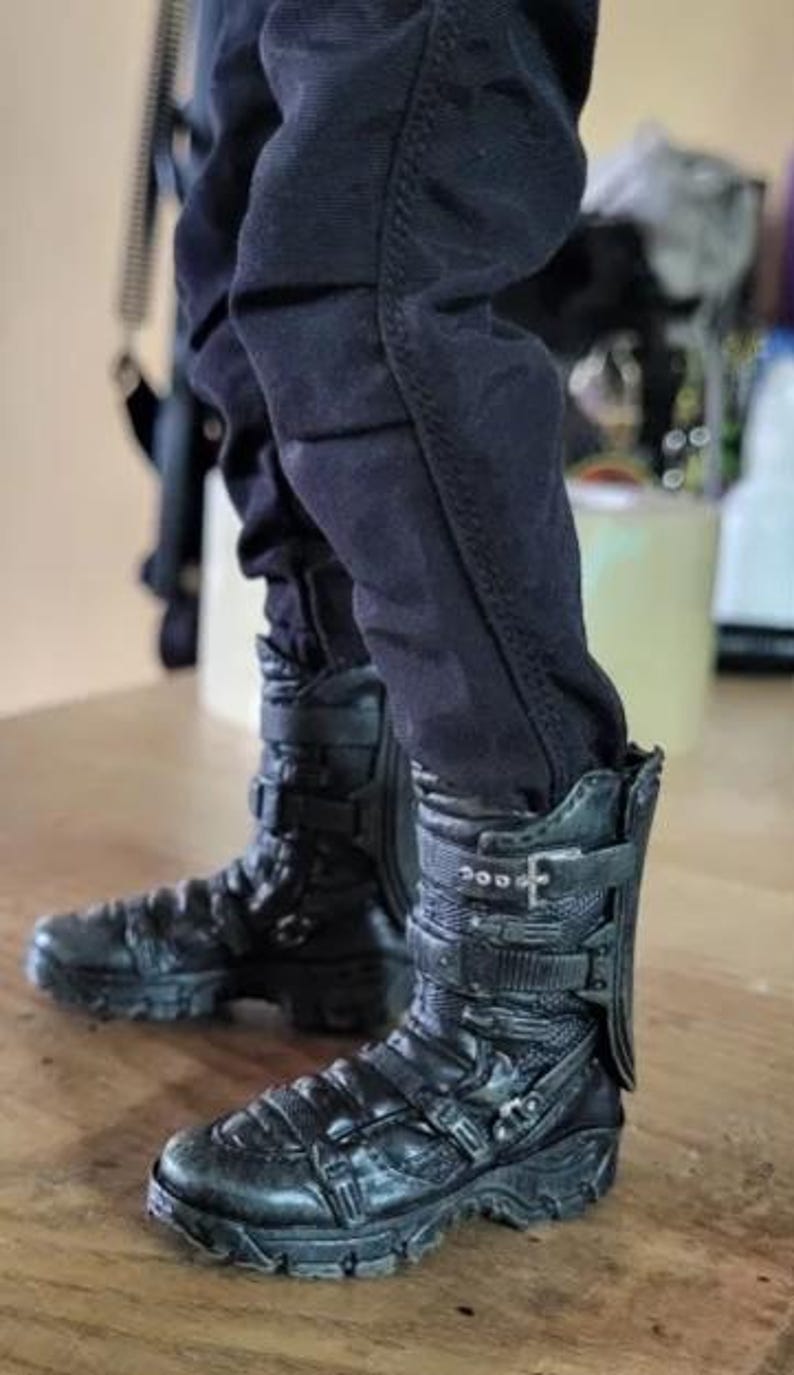 May include: Black combat boots with multiple buckles and straps. The boots have a rugged, textured appearance and are worn with black pants. The boots appear to be made of leather or a similar material, with visible stitching and detailing.