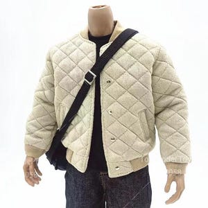 May include: A beige quilted bomber jacket with a ribbed collar and cuffs, featuring a front snap button closure and two side pockets. A black crossbody bag with a silver buckle strap is worn over the jacket, paired with a black shirt and dark jeans.