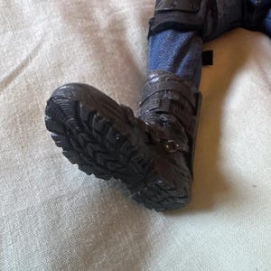 May include: Close-up of a black boot with a rugged sole. The boot is part of a figure wearing blue jeans and protective gear on the knee. The boot has multiple straps and buckles.