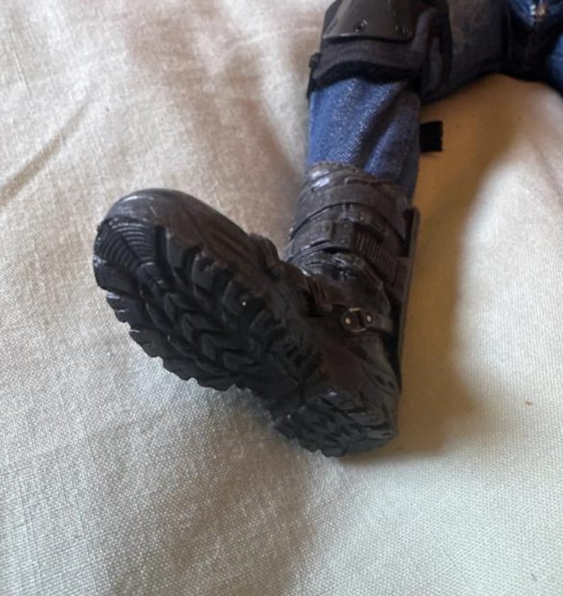 May include: Close-up of a black boot with a rugged sole. The boot is part of a figure wearing blue jeans and protective gear on the knee. The boot has multiple straps and buckles.