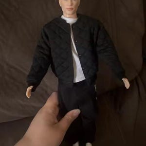 May include: A doll with brown hair styled in a top knot, wearing a black quilted bomber jacket over a white t-shirt, paired with black trousers and white trainers. The doll has a neutral facial expression and is standing in an upright position.