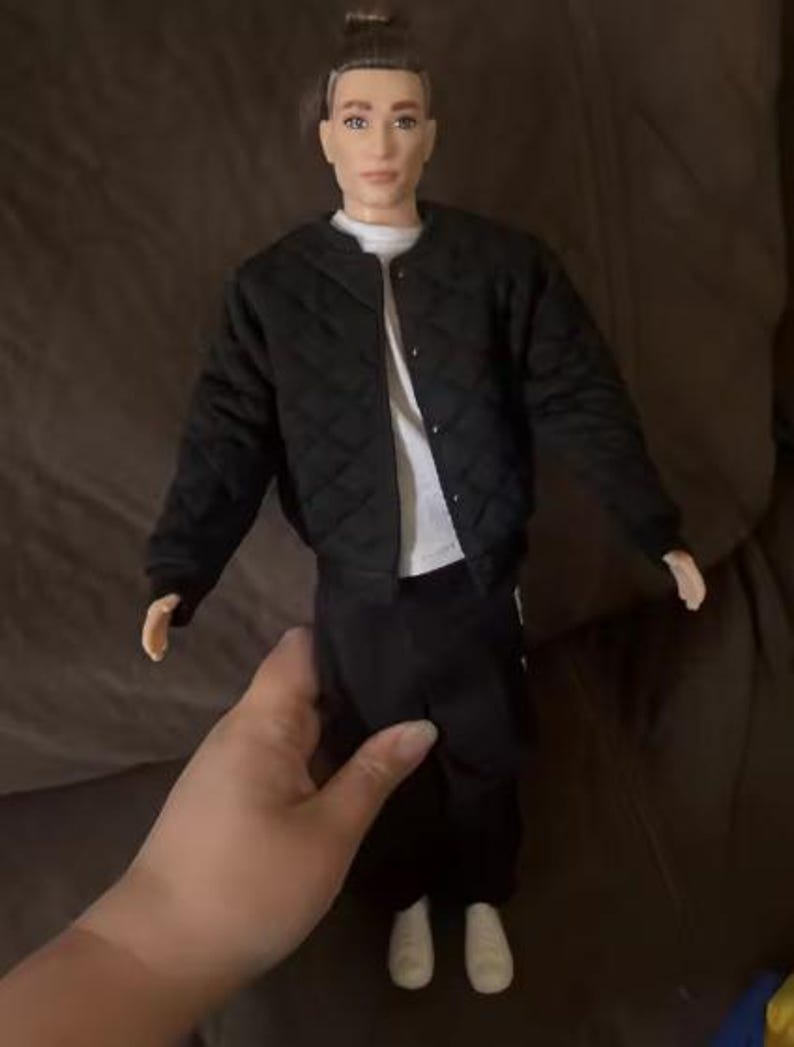 May include: A doll with brown hair styled in a top knot, wearing a black quilted bomber jacket over a white t-shirt, paired with black trousers and white trainers. The doll has a neutral facial expression and is standing in an upright position.
