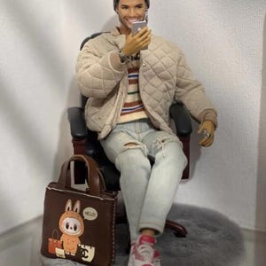 May include: A male fashion doll wearing a grey beanie, a beige quilted bomber jacket, a striped sweater, and light blue distressed jeans. The doll holds a small silver smartphone and sits on a black chair next to a brown tote bag with a cartoon graphic.